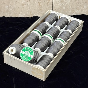 May include: A rectangular cardboard box containing twelve spools of dark gray polyamide machine twist thread. Each spool features a white end cap with the number 133 and a green label printed with the text SILKE UTICA THREADS.