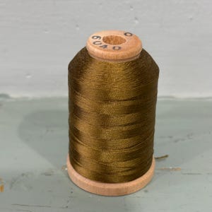 May include: A spool of dark olive green sewing thread wound onto a light-colored wooden core. The top of the wooden spool features the printed black text 6040. The thread has a smooth, slightly lustrous finish and is tightly wrapped.