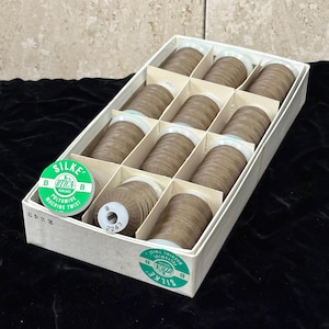 May include: A white cardboard box containing eleven spools of light brown Silke Utica Threads polyamide machine twist sewing thread. Each spool features a white plastic core and a green label with the text Silke Utica Threads.