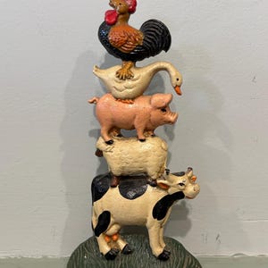 May include: A vertical cast metal figurine featuring a stack of farm animals standing on a green base. From bottom to top, the stack includes a black and white cow, a cream-colored sheep, a pink pig, a white goose, and a rooster with red and black details.