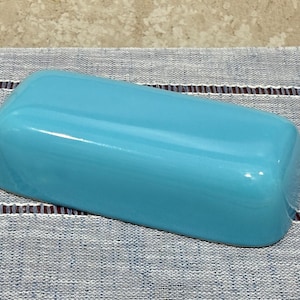 May include: A rectangular, solid turquoise butter dish cover with a smooth, glossy finish and rounded edges. The object has a simple, clean design with no visible patterns, textures, or markings on its exterior surface.