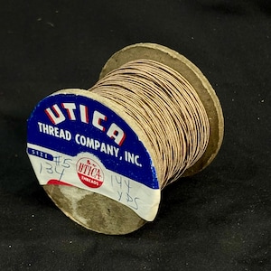 May include: A spool of tan thread from Utica Thread Company, Inc. The blue and white label features the text UTICA THREAD COMPANY, INC., size #5, and 144 YDS handwritten in black ink on the bottom white section of the label.