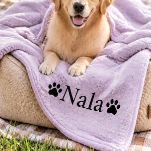 Peut inclure: Un chien golden retriever reposant sur un panier beige recouvert d'une couverture en peluche lavande douce. La couverture porte le nom Nala brodé en lettres noires, encadré par deux icônes d'empreintes de pattes noires de chaque côté.