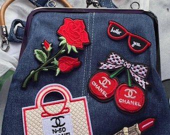 Op de afbeelding: Een donkerblauwe spijkerstof handtas met een metalen frame en sluiting, versierd met geborduurde patches waaronder een rode roos, een rode zonnebril met de tekst hello you, een geblokte strik met twee rode Chanel-kersen, een Chanel N-50 tas en een lippenstift.