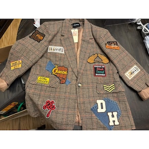 Op de afbeelding: Een bruine blazer met ruitpatroon en diverse kleurrijke geborduurde patches met de teksten Blessed, Queen, Goal Digger, Brown Suga Babe, There Is Nothing Basic, Minding My Black Owned Business, Love The Skin You Are In en DH.