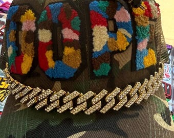 Op de afbeelding: Een camouflage baseballpet met het woord DOPE in veelkleurige, getextureerde, pluizige letters op de voorkant. Een goudkleurige ketting met doorzichtige strass-steentjes is aan de klep bevestigd en een zwarte kralenbedel zit bovenop.