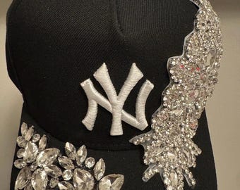 Op de afbeelding: Een zwarte honkbalpet met een wit geborduurd New York Yankees-logo op de voorkant. De pet is versierd met twee afzonderlijke secties van heldere, gefacetteerde kristallen decoraties op de zij- en voorpanelen.