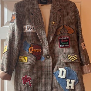 May include: A brown plaid blazer featuring various embroidered patches including text that reads Blessed, Queen, Goal Digger, Minding My Black Owned Business, and DH, along with a gold elephant pin and a white shirt underneath.