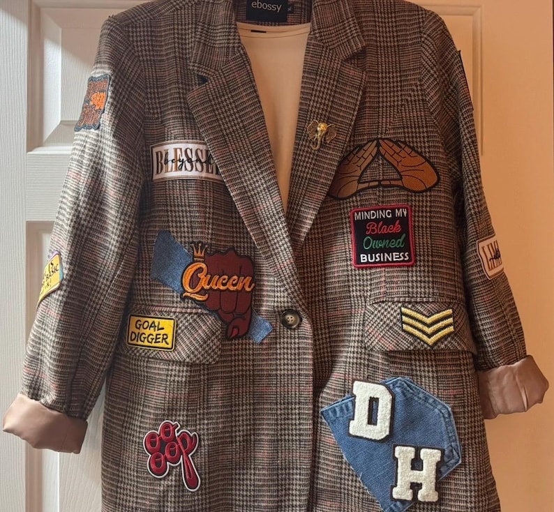 Op de afbeelding: Een bruine blazer met ruitpatroon voorzien van diverse geborduurde patches met teksten als Blessed, Queen, Goal Digger, Minding My Black Owned Business en DH, inclusief een gouden olifantspeld en een wit hemd eronder.