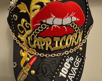 Op de afbeelding: Een zwarte honkbalpet met een geborduurd rood lipontwerp, gouden filigraan en het woord CAPRICORN. De pet is versierd met een gouden ketting, een veiligheidsspeld met witte kralen, een lippenstifticoon en een zwarte patch met 100% SAVAGE.