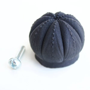 May include: A matte black cabinet knob with a rounded, segmented, pumpkin-like shape featuring small, raised dots along each vertical ridge. The knob is accompanied by a single silver-colored metal machine screw with a flat head and threaded shaft.