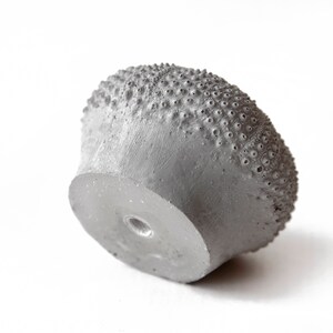 May include: A gray concrete knob with a textured surface resembling a sea urchin. The knob has a small hole in the center.
