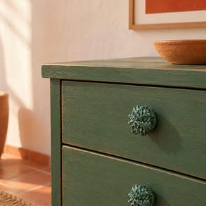 May include: A dark green wooden dresser featuring two drawers with textured, acorn-shaped knobs in a matching green finish. The dresser has a visible wood grain texture and a matte appearance, providing a functional storage unit for a room.