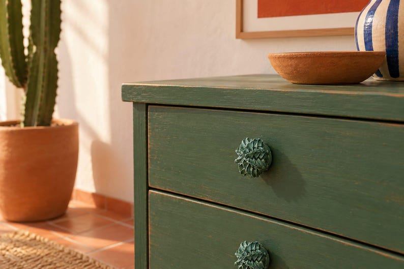 May include: A dark green wooden dresser featuring two drawers with textured, acorn-shaped knobs in a matching green finish. The dresser has a visible wood grain texture and a matte appearance, providing a functional storage unit for a room.