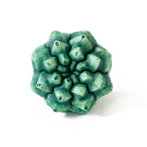 May include: A sculptural object shaped like a succulent plant with a geometric, multi-layered structure. The surface features a textured, matte finish in varying shades of deep forest green and lighter sage green, creating a realistic botanical appearance.