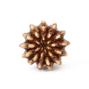 May include: A circular, star-shaped decorative knob with a textured, spiky surface. The object features a metallic bronze finish with lighter, cream-colored highlights on the tips of the spikes, creating a dimensional, radial pattern.