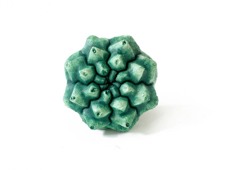 May include: A sculptural object shaped like a succulent plant with a geometric, multi-layered structure. The surface features a textured, matte finish in varying shades of deep forest green and lighter sage green, creating a realistic botanical appearance.