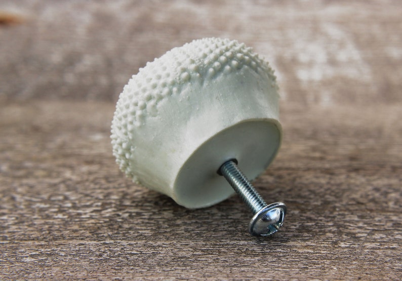 May include: White ceramic cabinet knob with a textured surface resembling a sea urchin. The knob has a silver screw.