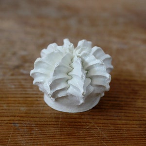 May include: A small, matte white ceramic sculpture featuring a rounded, multi-layered structure with ruffled, petal-like ridges radiating from the center. The object has a solid, flat circular base and a textured, organic appearance.