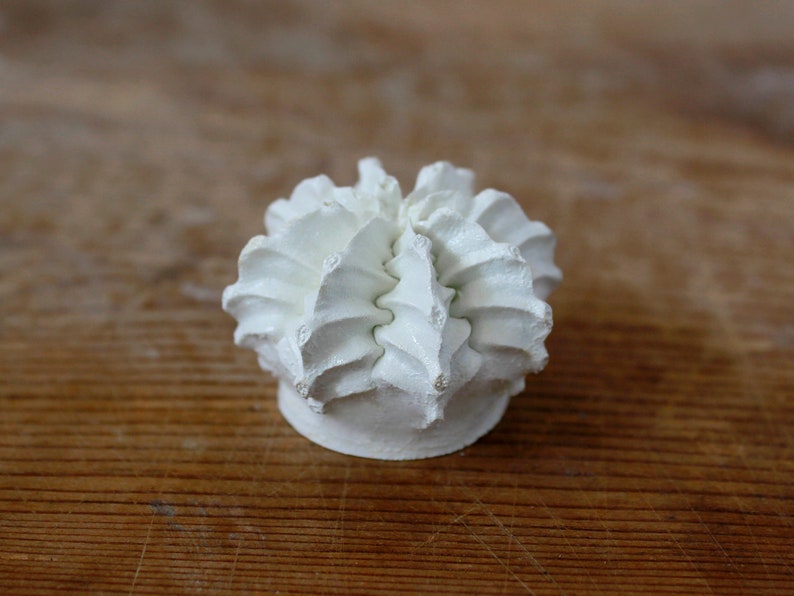May include: A small, matte white ceramic sculpture featuring a rounded, multi-layered structure with ruffled, petal-like ridges radiating from the center. The object has a solid, flat circular base and a textured, organic appearance.