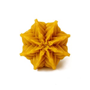 May include: A golden yellow wax object shaped like a six-pointed star or flower with intricate, layered, and pointed petals radiating from a central point. The surface has a smooth, matte texture with defined ridges and deep, symmetrical folds.