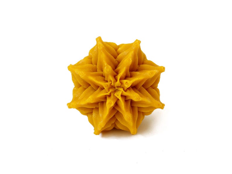 May include: A golden yellow wax object shaped like a six-pointed star or flower with intricate, layered, and pointed petals radiating from a central point. The surface has a smooth, matte texture with defined ridges and deep, symmetrical folds.