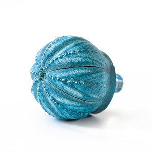 May include: A round, teal-coloured cabinet knob with a textured, ribbed surface resembling a sea urchin. The finish features a distressed, weathered appearance with lighter blue highlights along the raised ridges and small, dotted details.