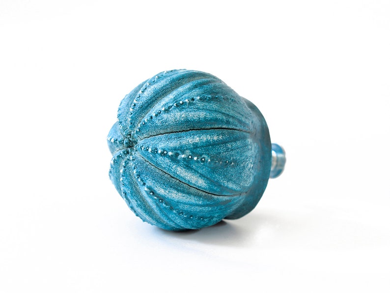 May include: A round, teal-coloured cabinet knob with a textured, ribbed surface resembling a sea urchin. The finish features a distressed, weathered appearance with lighter blue highlights along the raised ridges and small, dotted details.