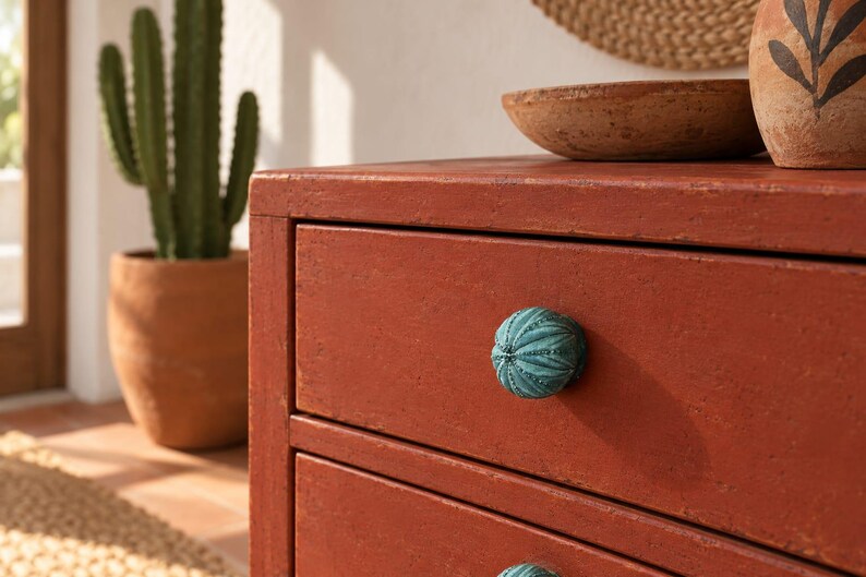May include: A round, textured drawer knob in a teal color with a ribbed, sea urchin-like pattern. The knob is attached to a terracotta-colored wooden drawer front, which features a matte finish and visible wood grain texture on the surface.