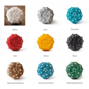 May include: Nine sea urchin shells painted in various colors, including white, gray, turquoise, red, yellow, black, brown, blue, and green. The shells are arranged in a grid pattern on a white background.