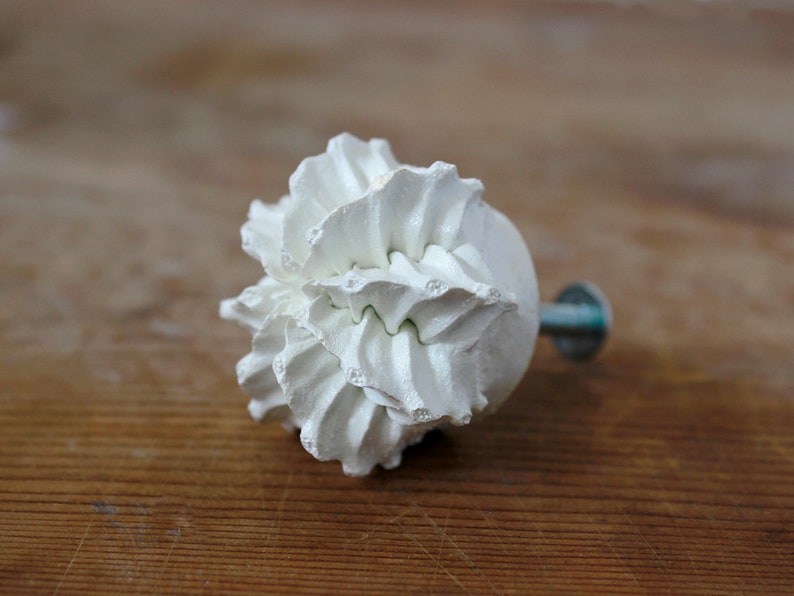May include: A white cabinet knob shaped like a ruffled flower or a dollop of whipped cream. The front features intricate, layered, and textured petals radiating from the center, while a metal screw extends from the back for mounting purposes.