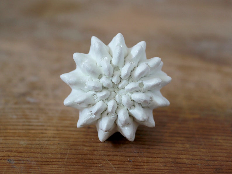 May include: A white ceramic knob with a textured, multi-layered, star-shaped design. The surface features numerous pointed, petal-like protrusions radiating from the center, creating a dense, geometric, and three-dimensional floral appearance.