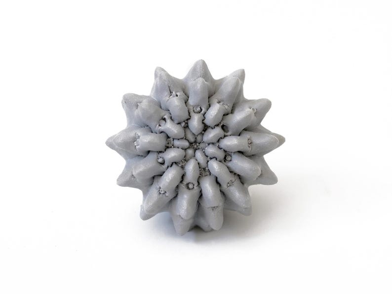May include: A light gray, spherical object with a complex, repeating geometric pattern of pointed, star-like protrusions radiating from the center. The surface features a dense, textured arrangement of small, interlocking spikes in a uniform color.