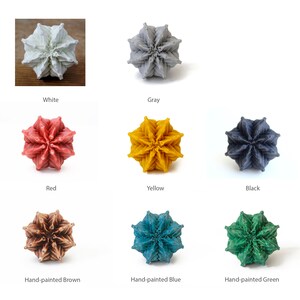 May include: A collection of eight geometric, star-shaped objects arranged in a grid. The items are available in solid white, gray, red, yellow, and black, as well as hand-painted brown, blue, and green finishes, each featuring a complex, folded texture.