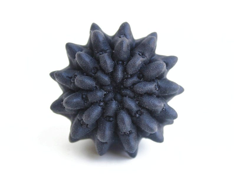 May include: A dark charcoal gray, star-shaped object with a textured, matte surface. The item features numerous rounded, protruding points radiating from a central core, creating a dense, multi-layered, and symmetrical floral-like structure.
