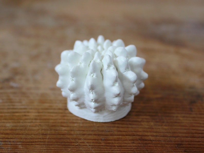 May include: A small, white, spherical object with a textured, bumpy surface resembling a cactus or a sea urchin. The item features numerous rounded, protruding points across its top and sides, resting on a flat, circular base.