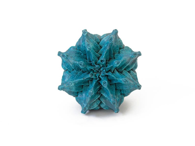 May include: A teal-colored, multi-faceted geometric object with a star-like, symmetrical structure. The surface features a textured, matte finish with deep blue and green tones, composed of numerous pointed, triangular segments radiating outward.