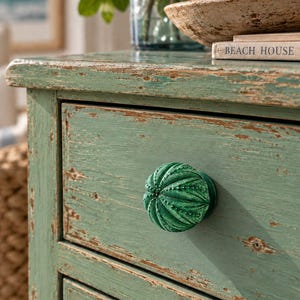 May include: A round, dark green cabinet knob with a textured, ribbed surface resembling a sea urchin shell. The knob is attached to a light green wooden drawer front that features a distressed, weathered finish with visible wood grain.