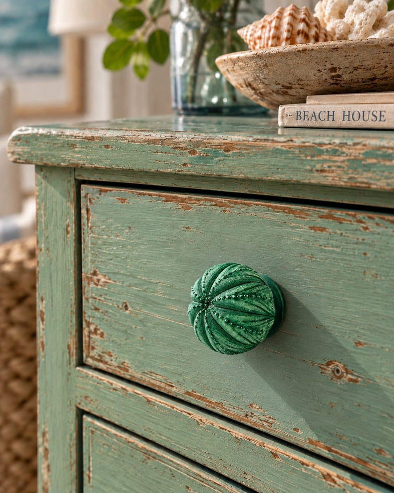 May include: A round, dark green cabinet knob with a textured, ribbed surface resembling a sea urchin shell. The knob is attached to a light green wooden drawer front that features a distressed, weathered finish with visible wood grain.