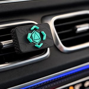 May include: A rectangular black car air freshener vent clip featuring a topographic map pattern on its surface. The center displays a raised teal geometric emblem with a triangle and four surrounding arrow-shaped accents.