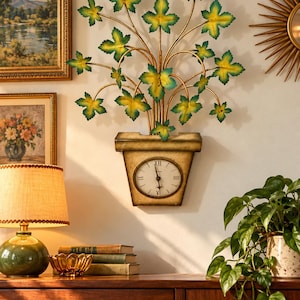 May include: A wall-mounted clock featuring a gold-toned planter base with a round white face and black Roman numerals. Extending upward from the planter are metal stems with green and yellow leaves, creating a botanical wall sculpture.
