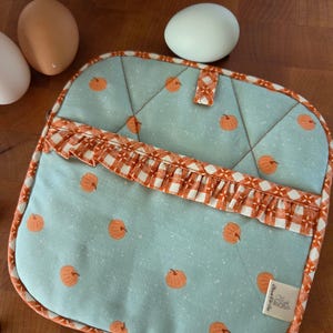 Puede incluir: Un agarrador de cocina acolchado cuadrado de tela azul claro con un patrón repetitivo de calabazas naranjas. Incluye un volante a cuadros naranjas y blancos, un ribete a juego y una pequeña etiqueta de tela blanca.