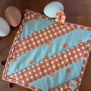 Puede incluir: Un agarrador de cocina cuadrado y acolchado con franjas diagonales alternas de tela azul claro con estampado de pequeñas calabazas naranjas y tela de cuadros vichy naranja y blanco. Incluye una trabilla a juego y una etiqueta blanca.