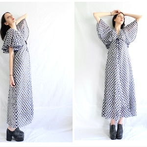 May include: A white maxi dress with a blue polka dot pattern and bell sleeves. The dress has a V-neckline and a tie at the waist. The model is wearing black platform shoes.