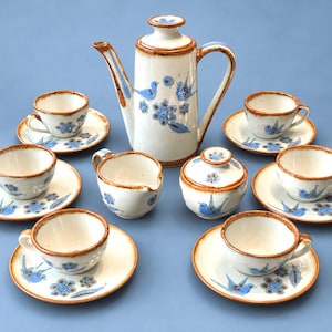 May include: A ceramic tea set featuring a tall teapot, a creamer, a lidded sugar bowl, and six cups with matching saucers. Each piece is off-white with hand-painted blue birds and floral motifs, finished with a brown glazed rim.