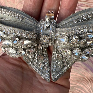 May include: A silver bow with a crystal embellishment. The bow has a silver clasp in the center.