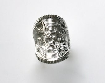 May include: A silver ring with a circular design that resembles a tree trunk. The ring has a textured surface and a slightly raised edge.