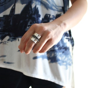 May include: A silver and black wide band ring with a textured design. The ring is worn on the index finger of a person's hand.