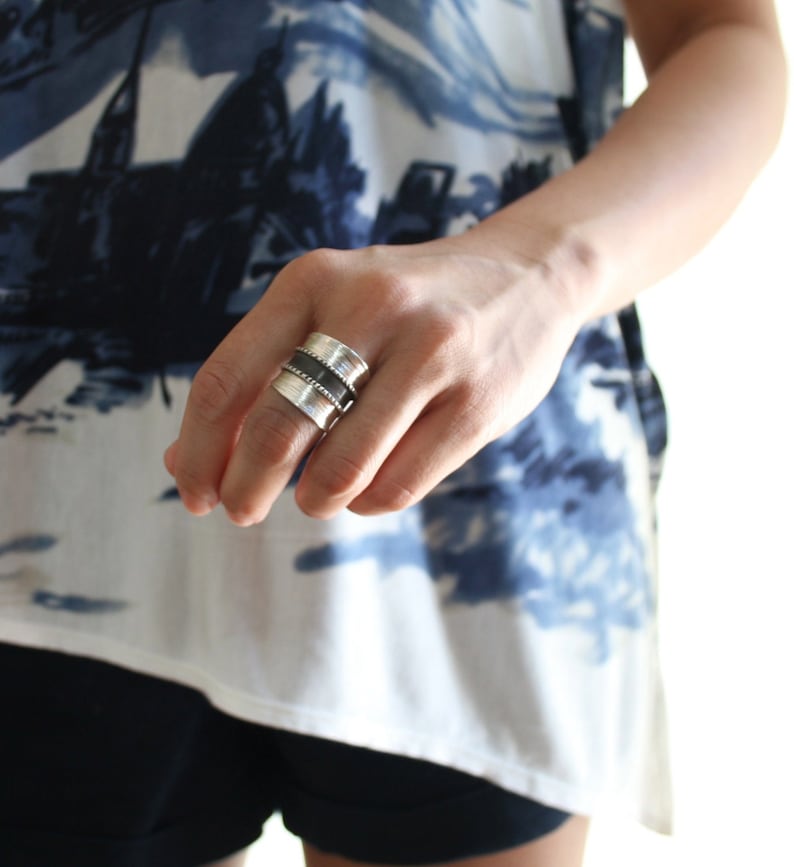 May include: A silver and black wide band ring with a textured design. The ring is worn on the index finger of a person's hand.
