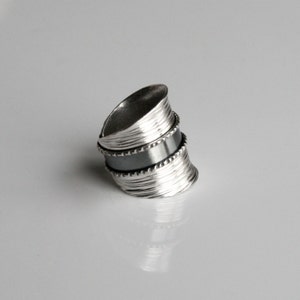 May include: A silver ring with a textured, layered design. The ring has a black band in the middle.